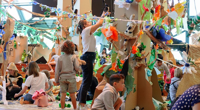 A woman reaches above her head to hand artwork on the wall of a room that is filled with children's crafted arts. There are five children throughout the room interacting with the art and creating. 